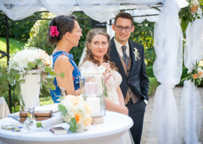 Bei einer Hochzeitszeremonie stehen Braut, Bräutigam und Rednerin Mandy Römer beim Kerzenritual nebeneinander.