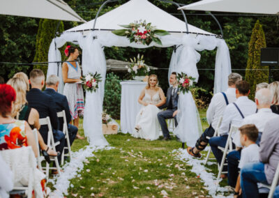Eine Hochzeitszeremonie in einem Garten mit Brautpaar, Rednerin Mandy Römer und Gästen.