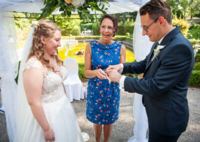 Eine Braut und ein Bräutigam tauschen ihre Eheringe neben Rednerin Mandy Römer unter einem Pavillon aus.